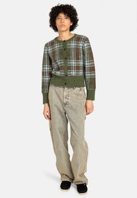 Zeleni karo kardigan s gumbima sprijeda, rebrastim manžetama i rubom, uparen s širokim svijetlim trapericama i crnom obućom.