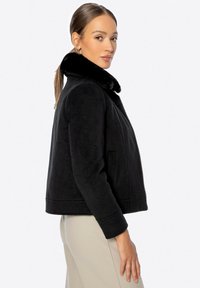 Chaqueta negra de piel sintética con cuello de piel, diseño corto, mangas largas, dos bolsillos frontales y una textura suave. Presenta una silueta limpia y minimalista.