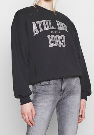 Osoba nosząca czarną bluzę z napisem "ATHL. DEPT 1983 AMERICA" i jasnoszare dżinsy, stojąca na jednolitym tle.