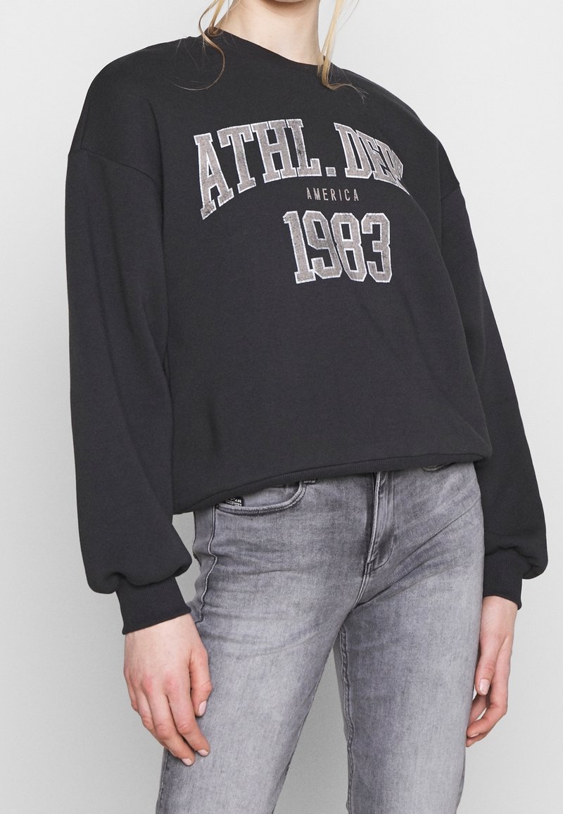 Personne portant un sweat-shirt noir avec le texte "ATHL. DEPT 1983 AMERICA" et un jean gris clair, debout devant un fond uni.
