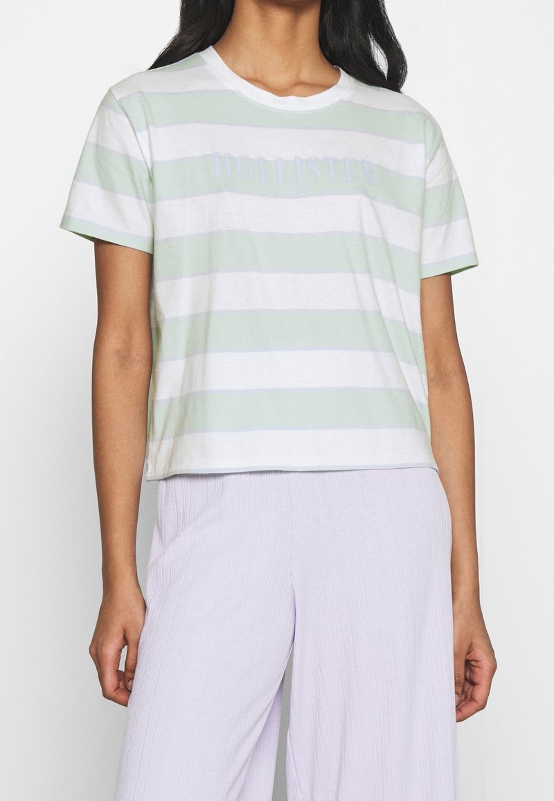 Tričko s krátkým rukávem a kulatým výstřihem v světle zelené barvě s bílými pruhy, se vyšitým logem značky v světle modré barvě na hrudi.