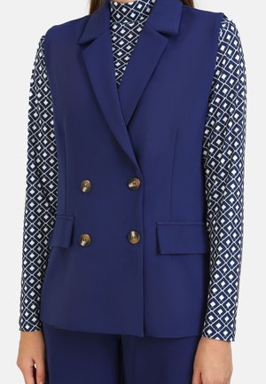 Gilet blu navy doppiopetto realizzato in tessuto morbido, con bottoni decorativi dorati, tasche laterali, indossato su una camicia a maniche lunghe con motivo.
