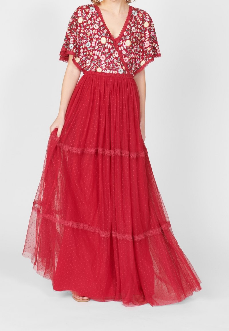 Femme portant une robe rouge longue jusqu'au sol avec un corsage brodé de fleurs et une jupe transparente à pois, tenant le tissu d'une main.