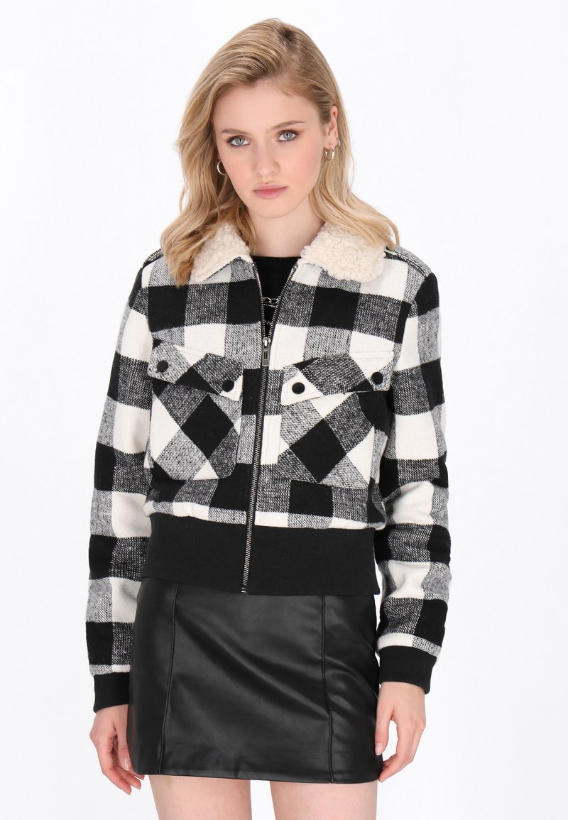 Giacca a scacchi bianco e nero con colletto sherpa, chiusura con zip e due tasche frontali, abbinata a una mini gonna in pelle nera.