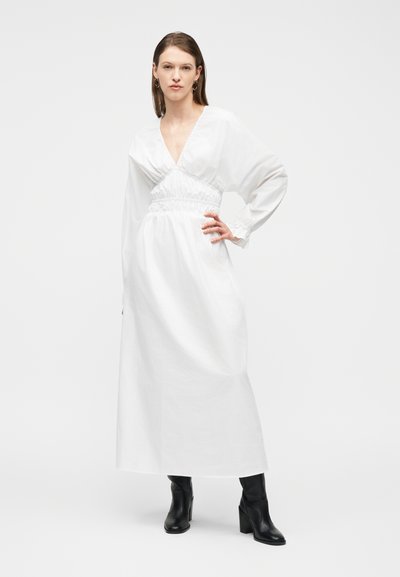 Γυναίκα με ίσια καστανά μαλλιά, φοράει μακρύ λευκό φόρεμα με σφηκοφωλιά στη μέση και μαύρες μπότες με τακούνι, στέκεται με το ένα χέρι στη μέση.