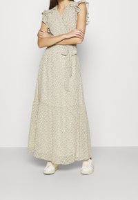 Vaaleanbeige kukallinen maksimekko, jossa on röyhelöhihat, V-pääntie, nauhavyötärö ja kerroksittainen helma. Yhdistetty valkoisiin tennareihin kontrastia varten.