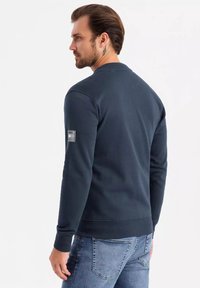 Felpa blu navy con maniche lunghe, polsini e orlo a coste, e una piccola patch sul braccio sinistro, indossata con jeans di denim.