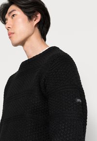Maglione nero a maglia con motivi texturizzati, collo a giro, e un piccolo badge con logo sulla manica sinistra. Tessuto morbido e spesso.