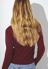 Long, wavy blonde hair viewed from the back. The person wears a burgundy long-sleeve top and light blue denim jeans. Minimalist setting.