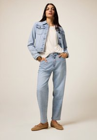 Chaqueta de mezclilla azul claro con un diseño recortado, combinada con jeans azul claro que presentan un patrón de puntos sutil. Unos zapatos marrones casuales completan el conjunto.