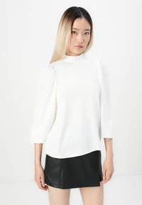 Young woman with straight ombre hair wearing a white long-sleeve blouse and a black leather mini skirt, standing against a plain white background.
