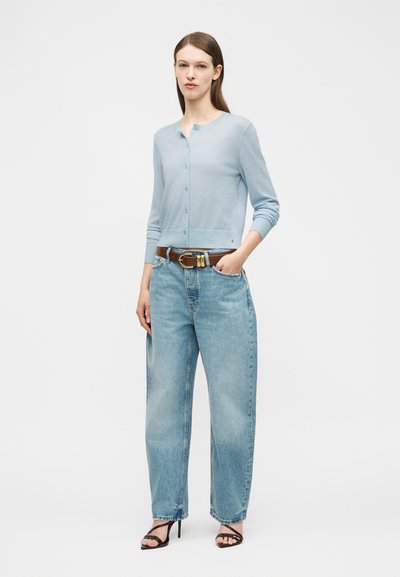Vrouw staat, draagt een lichtblauwe gebuttonde cardigan, lichtblauwe losse spijkerbroek met een bruine riem en zwarte sandalen met hak, hand in zak.