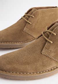 Brune ruskindssko med en rund tå, der har beige snørebånd og hvide syninger langs sålen. Glat tekstur og casual design.