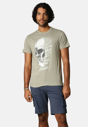 Olijfgroen T-shirt met een grote schedelafbeelding en tekstprint. Korte mouwen, ronde halslijn, zachte katoen stof. G gedragen met marineblauwe cargoshorts.