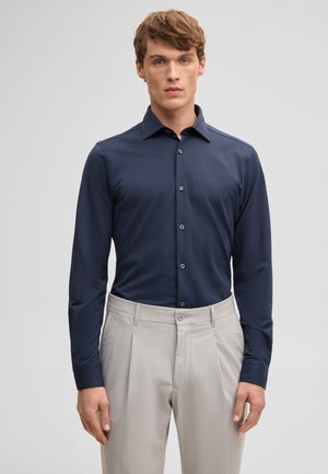 Mężczyzna w dopasowanej granatowej koszuli na guziki, włożonej w jasnobeżowe plisowane spodnie, stojący na jednolitym tle.