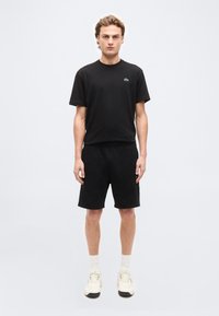 Νεαρός άνδρας στέκεται προς τα εμπρός φορώντας μαύρο T-shirt με μικρό λογότυπο, μαύρο σορτς, λευκές κάλτσες και λευκά αθλητικά παπούτσια σε απλό φόντο.