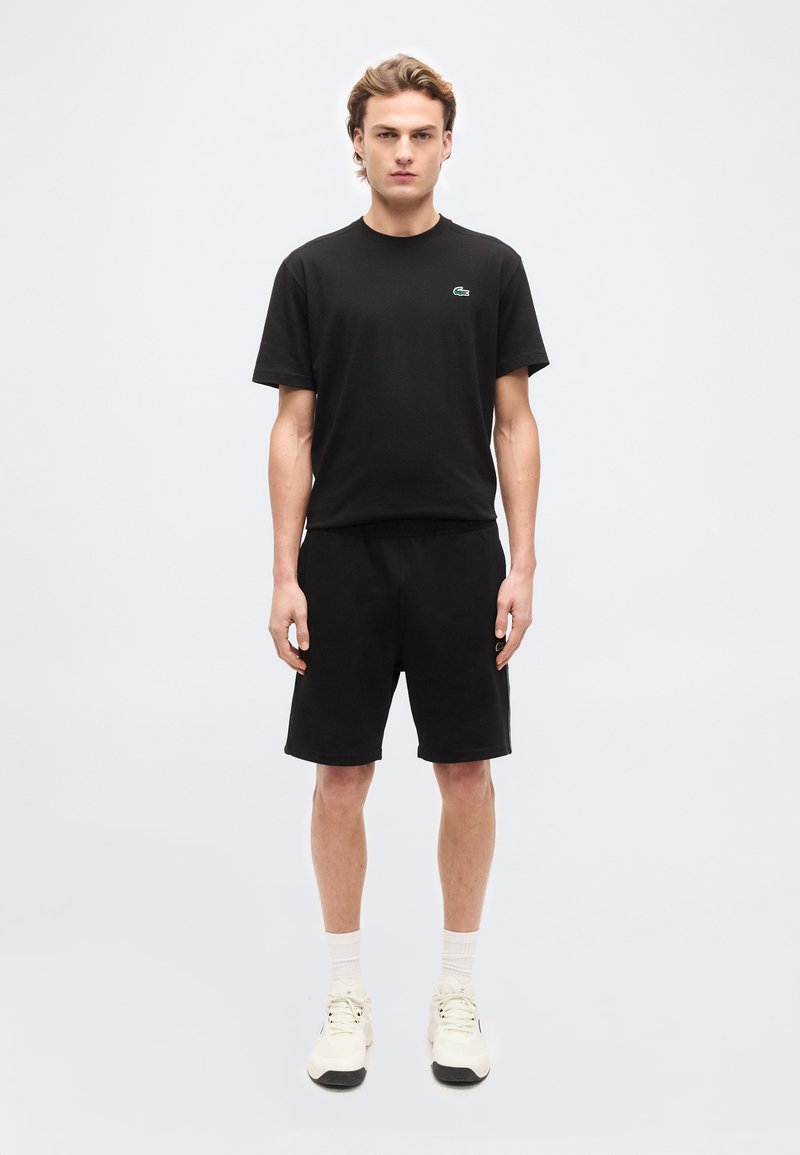 Νεαρός άνδρας στέκεται προς τα εμπρός φορώντας μαύρο T-shirt με μικρό λογότυπο, μαύρο σορτς, λευκές κάλτσες και λευκά αθλητικά παπούτσια σε απλό φόντο.