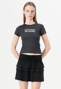 Sort svart t-skjorte med hvit tekst "CATCH FLIGHTS NOT FEELINGS"; kombinert med en svart, lagdelt mini skjørt med snøring i livet.