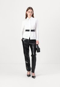 Camicia bianca a maniche lunghe con bottoni, abbinata a una cintura nera in vita, pantaloni lucidi neri a gamba dritta e tacchi neri.