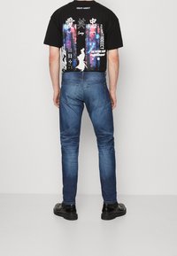 Musta graafinen t-paita, jossa monivärisiä painokuvioita takana, yhdistetty kapealahkeisiin sinisiin farkkuihin ja mustiin nilkkureihin.