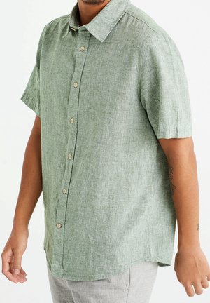 Uomo con camicia verde chiaro a maniche corte abbottonata, abbinata a pantaloni grigio chiaro, in piedi con le braccia leggermente piegate.