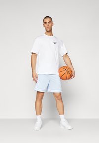 Hvit t-skjorte med lite logo, lyseblå perforerte basketballshorts med hvite detaljer, hvite joggesko, og holder en oransje basketball.