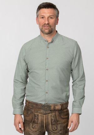 Mann trägt grünes kariertes Hemd und braune bestickte Lederhosen mit dekorativer Gürtelschnalle, steht vor einfachem Hintergrund.