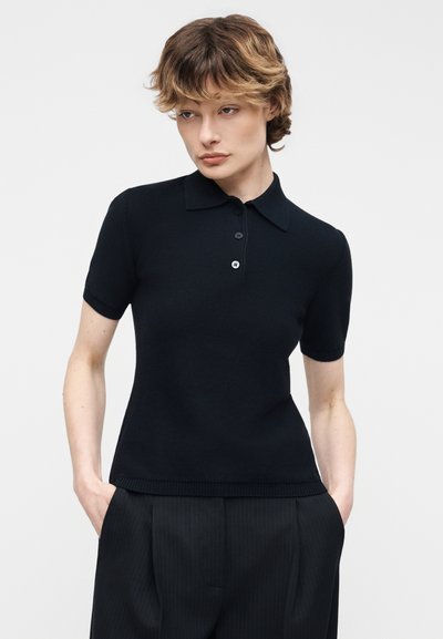 Γυναίκα με κοντά κυματιστά μαλλιά, φορώντας μαύρο πόλο μπλουζάκι με κοντά μανίκια και μαύρο παντελόνι με ρίγες, με τα χέρια στις τσέπες, κοιτάζοντας προς τα κάτω και αριστερά.