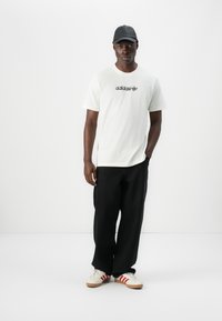 White cotton t-shirt with black Adidas logo, black wide-leg pants, and cream sneakers with red stripes, paired with a black cap.