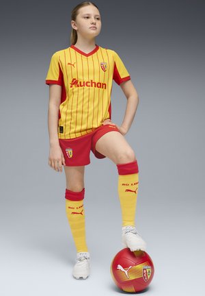 Ungdomsfotbollskit i gult och rött med vertikala ränder; inkluderar shorts, strumpor och en boll. Har Puma-logotyp och sponsorbranding.