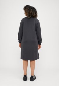 Grijze sweatdress met lange mouwen, geribbelde manchetten en studded accenten op de schouders. Gladde textuur, valt tot halverwege de kuit, gecombineerd met zwarte plateau-schoenen.