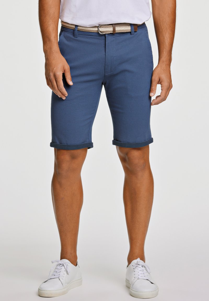 Shorts chino blu navy con orli risvoltati, realizzati in un tessuto testurizzato e dotati di una cintura intrecciata. Abbinati a sneakers bianche su uno sfondo neutro.