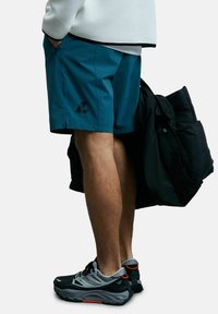 Shorts de sport teal avec un logo noir, associés à des chaussures noires avec des accents gris et orange. La texture semble lisse ; le design est simple.