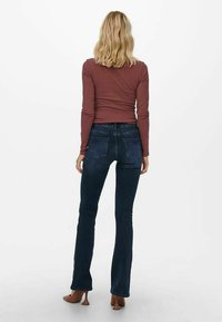 Jeans a zampa d'elefante di un blu scuro abbinati a un top aderente a maniche lunghe marrone. Il tessuto appare liscio con una sottile texture. La vista posteriore mostra i dettagli delle tasche.