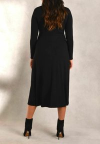 Musta pitkähihainen mekko, jossa on tiukka yläosa ja valuva halkio, valmistettu pehmeästä kankaasta. Malli käyttää mustia nilkkureita, joissa on korkea korko.