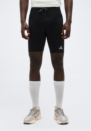 Moški, ki nosi črne atletske kratke hlače do sredine stegna z logotipom ACG, bele nogavice s zaokroženim robom ter bež in črne tekaške čevlje, stoji ob preprostem ozadju.