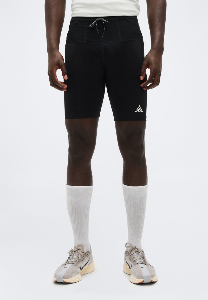 Άνδρας που φοράει μαύρα αθλητικά σορτς μήκους ως τη μέση μηρού με λογότυπο ACG, άσπρες κάλτσες και αθλητικά παπούτσια σε μπεζ και μαύρο, στέκεται μπροστά σε απλό φόντο.