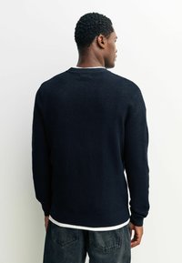 Marineblauer Strickpullover mit strukturiertem Muster, gerippten Bündchen und Kragen, über einem weißen Hemd getragen, von hinten betrachtet.