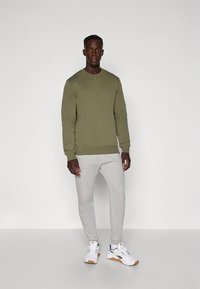 Felpa verde oliva con scollo rotondo, abbinata a pantaloni della tuta aderenti color grigio chiaro. Sneakers bianche con dettagli neri. Texture di tessuto liscia.