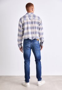 Camisa de cuadros en azul pálido y blanco con mangas largas, usada sobre unos vaqueros azules y zapatillas blancas. La camisa tiene un dobladillo curvado y yugo trasero.