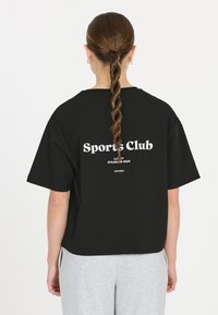 Sort oversized t-shirt med hvid tekst, "Sports Club," på ryggen. Korte ærmer og en afslappet pasform. Sammen med lysegrå sweatpants.