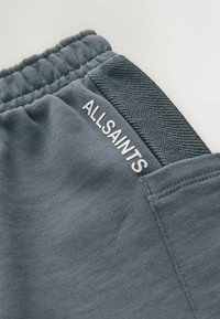 Grå atletikshorts med en tekstureret linning, der har mærkenavnet "ALLSAINTS" trykt i hvid på siden. Glat stoftekstur.