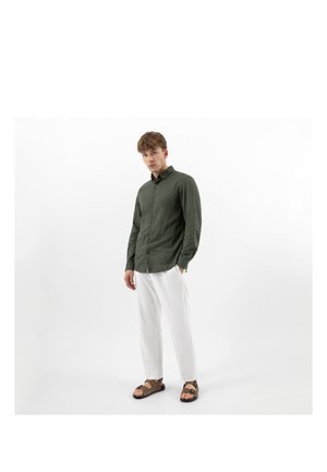 Giovane in piedi, indossa una maglia verde oliva a maniche lunghe, pantaloni bianchi e sandali marroni su sfondo bianco uniforme.