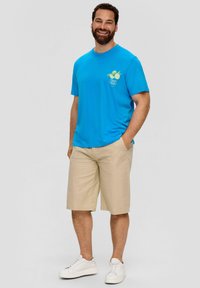 Blauwe katoenen t-shirt met een fruitafbeelding, gecombineerd met beige shorts. Het model draagt witte sneakers en laat een casual zomerset zien.