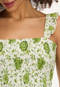 Weißes Kleid mit grünem Blumenmuster, das ein gerafftes Design und einen rüschenden Träger aufweist. Texturiertes Material verstärkt die visuelle Detailtiefe.