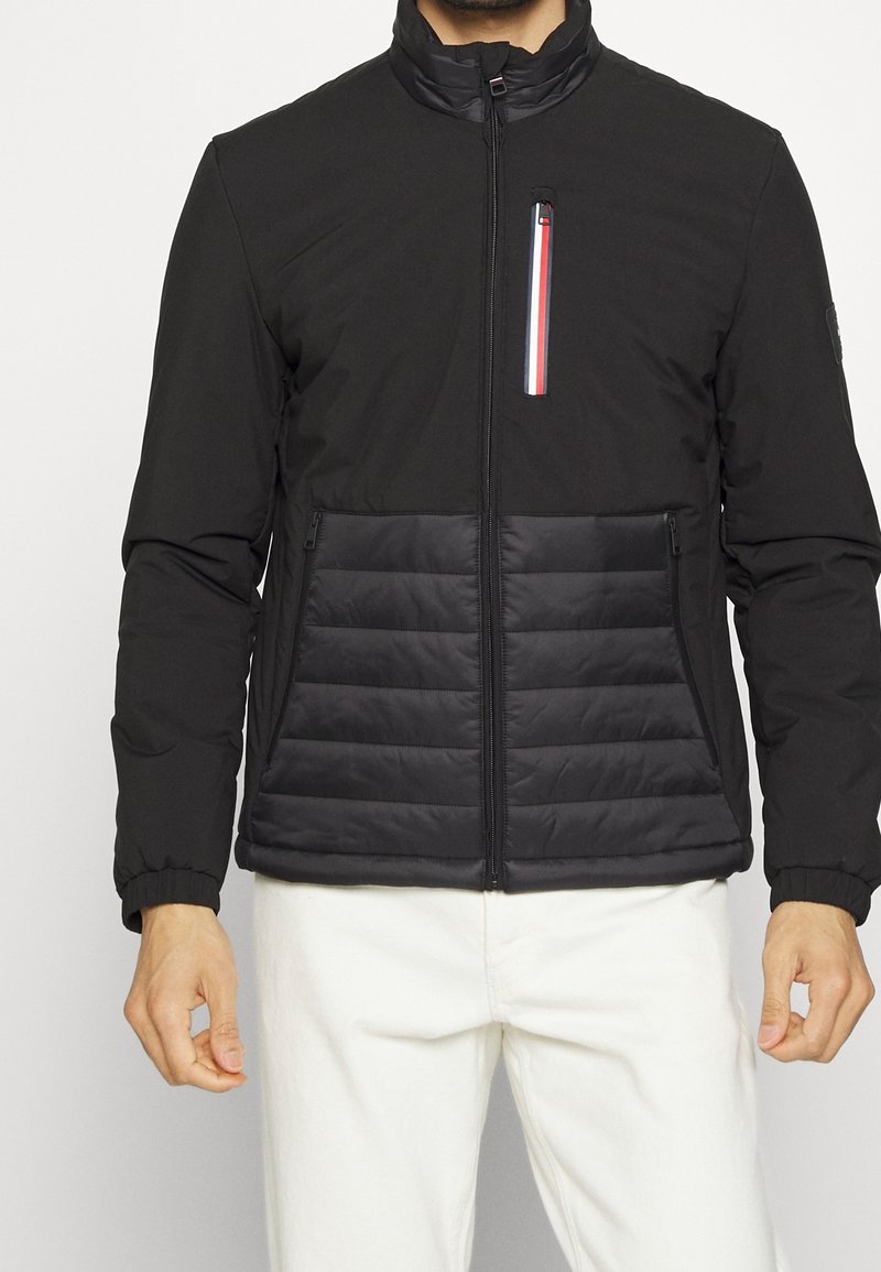 Black jacket with a high collar, front zipper and two side pockets. Features a quilted bottom panel and a pocket with red, white, and blue accents.