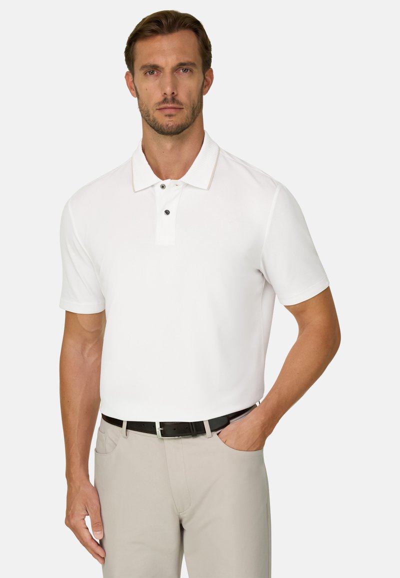 Homme portant un polo blanc à manches courtes, un pantalon beige et une ceinture noire, debout avec une main dans la poche devant un fond uni.