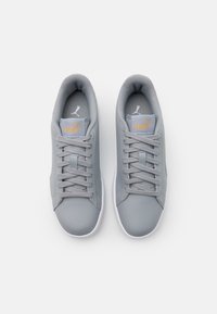 Šedé sportovní boty s hladkým syntetickým svrškem, plochými tkaničkami, polstrovaným límečkem a bílou gumovou podrážkou. Na jazyku je zlaté logo Puma.