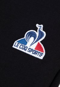 Svart tyg med en färgglad broderad logotyp som visar en stiliserad tupp och texten "LE COQ SPORTIF" i vitt och röda accenter.