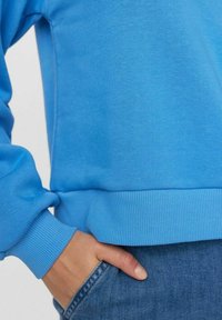 Šviesiai mėlynas džemperis su gumbuotais rankogaliais ir apačia, pasižymintis lygiu paviršiumi. Ranką įkišta į tamsiai mėlynų džinsų kišenę.